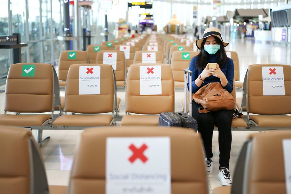 Young woman sits alone in airport at gate wearking mask and social distancing