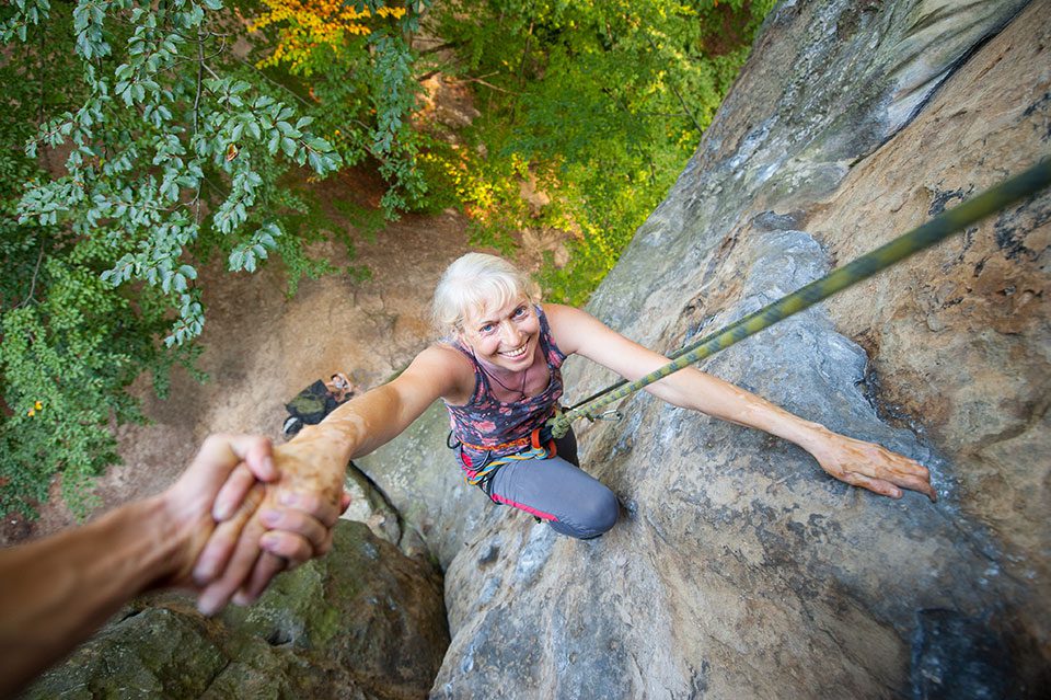 Older woman rock climbing smiles up at camera showing age-defying travel adventures