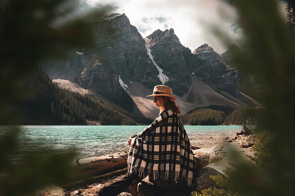 Older woman sits on a log wrapped in a blanket, mountains in the background