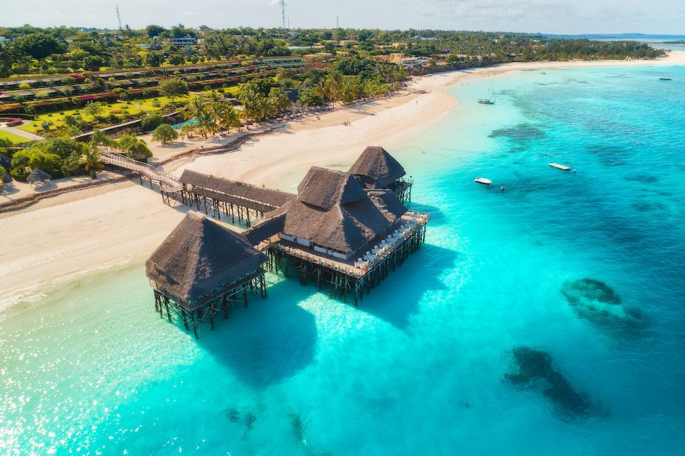 Aerial view of beautiful hotel on the water in ocean at sunset in summer. Zanzibar, Africa. Top view. Landscape with wooden hotel on the sea, azure water, sandy beach, green palm trees. Luxury resort