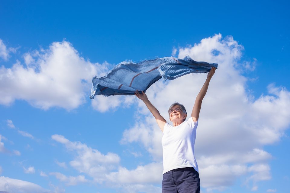 Happy mature elderly woman outdoors waving scarf in the wind. Smiling caucasian elderly woman with short hair wearing glasses enjoying freedom and vacation