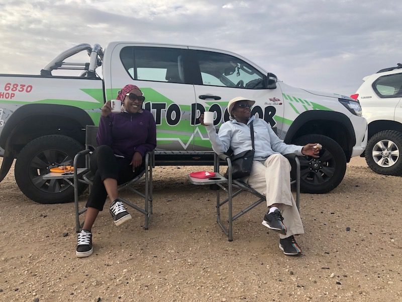 Two ladies enjoy breakfast next to a pickup truck after their hot air balloon safari experience in the Namib Desert