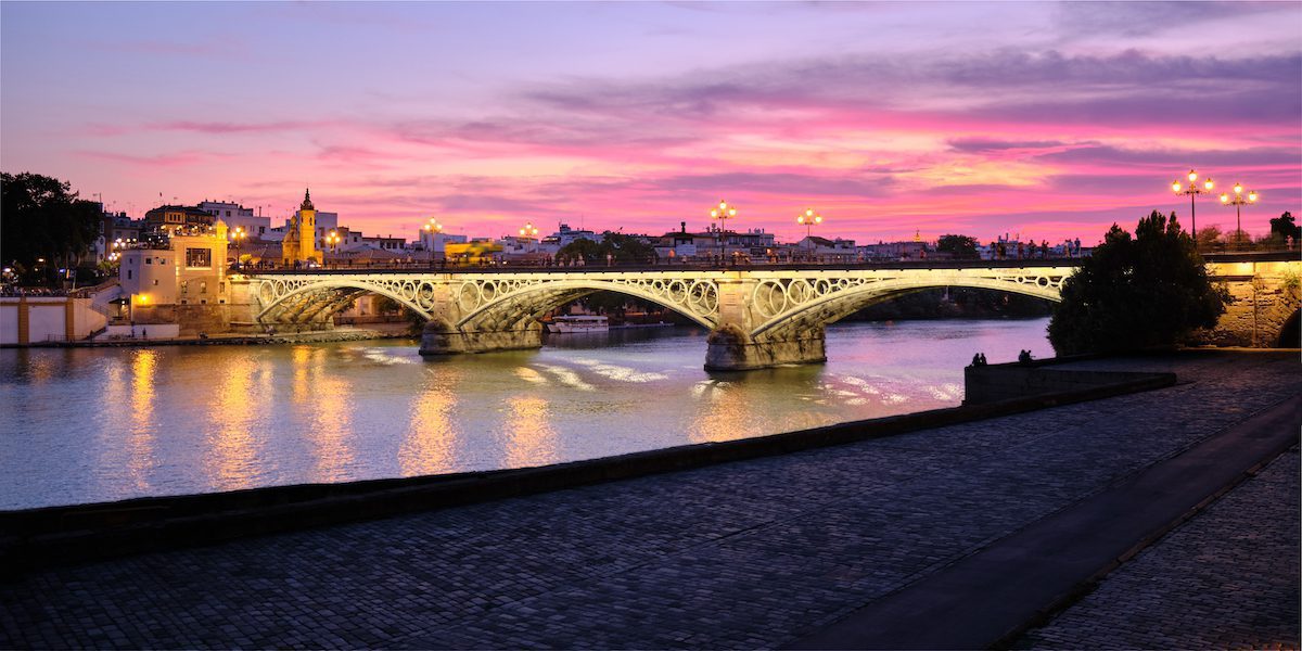 The Triana Bridge, Or Puente De Isabel II, Over The Guadalquivir River in Seville, Spain, At Dusk.