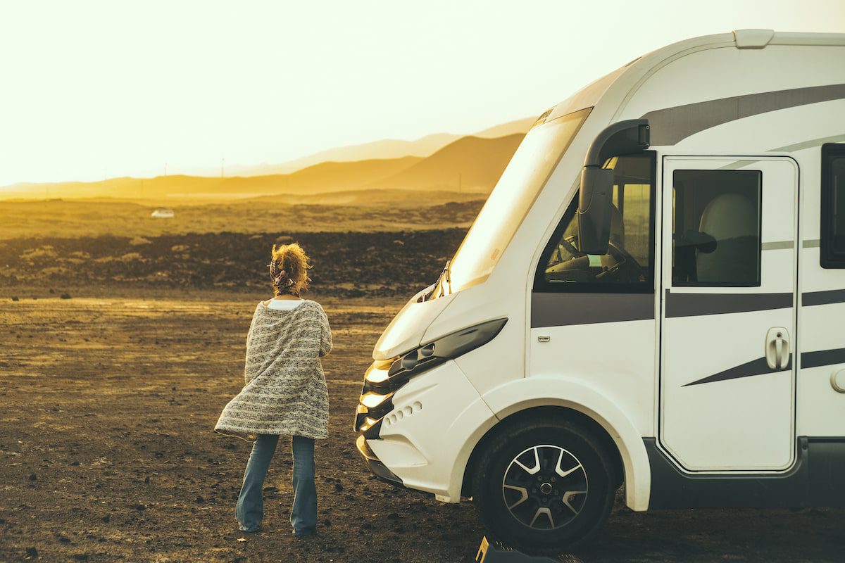 A woman stands beside her camper van RV at sunset in a field.