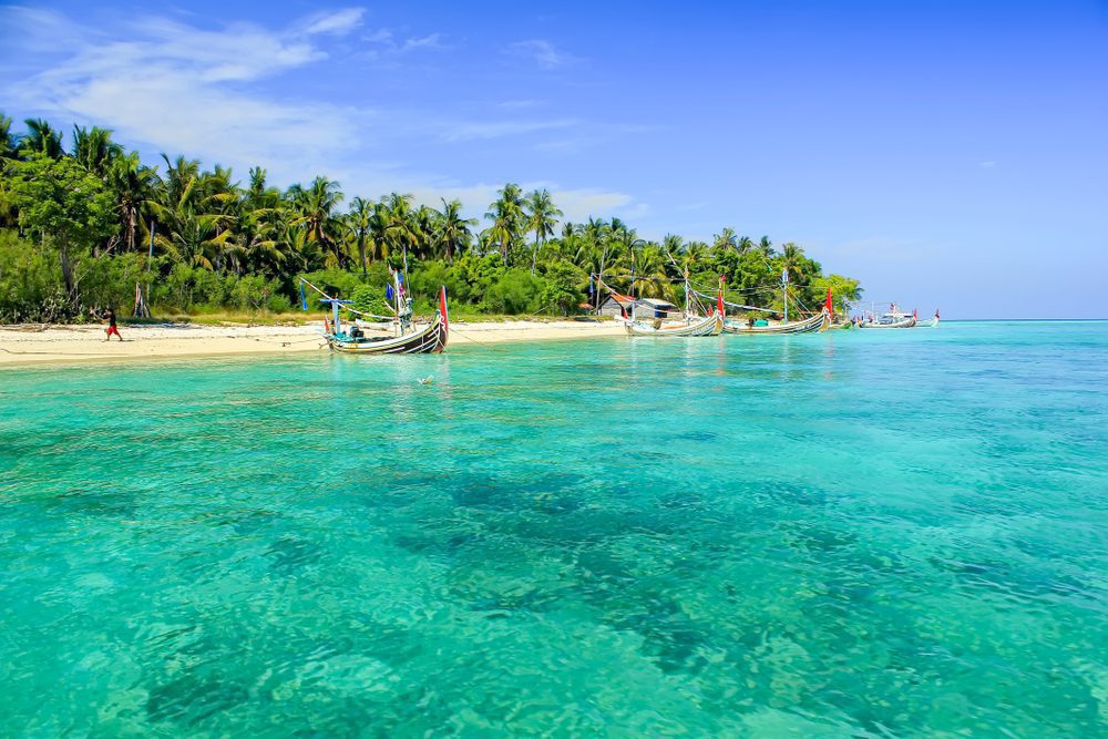 Crystal blue water with golden sand and green forest in the background. boats float in the shore.