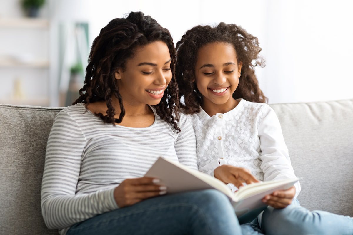 A mother reads a book with her daughter on the couch.