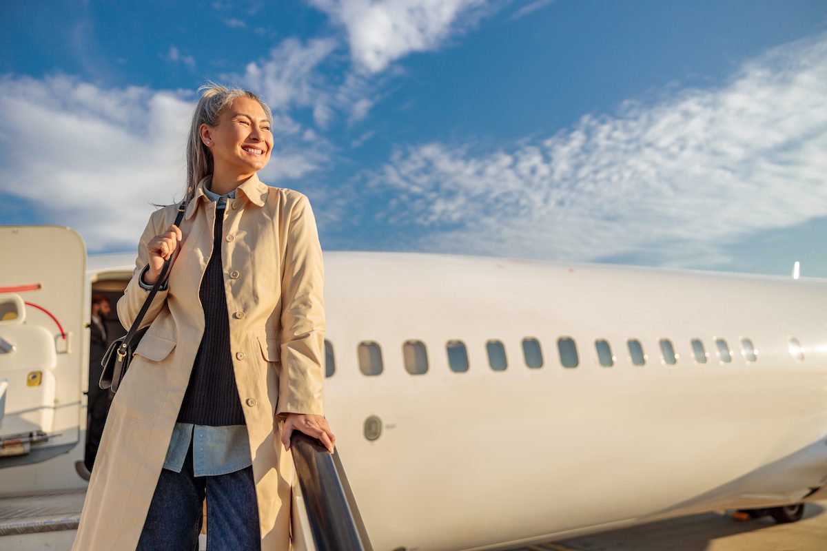 Joyful female traveler looking away and smiling while standing on airplane boarding stairs under blue cloudy sky