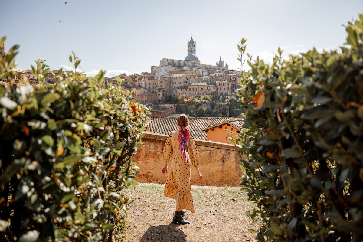 A woman in a long dress walks with Tuscan town of Siena, Italy in the background