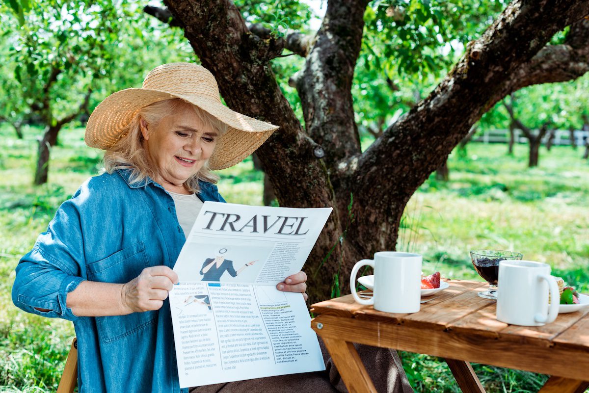 Senior woman in straw hat sitting near tree and reading travel newspaper after learning how to have trust in travel media